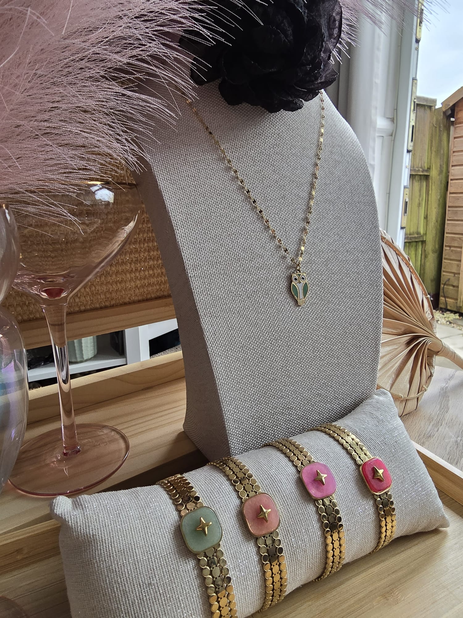 The Art of Shine Stella Polare bracelets in pastel pink and green, featuring gold-tone coin chains and a central North Star motif, displayed on a linen cushion.